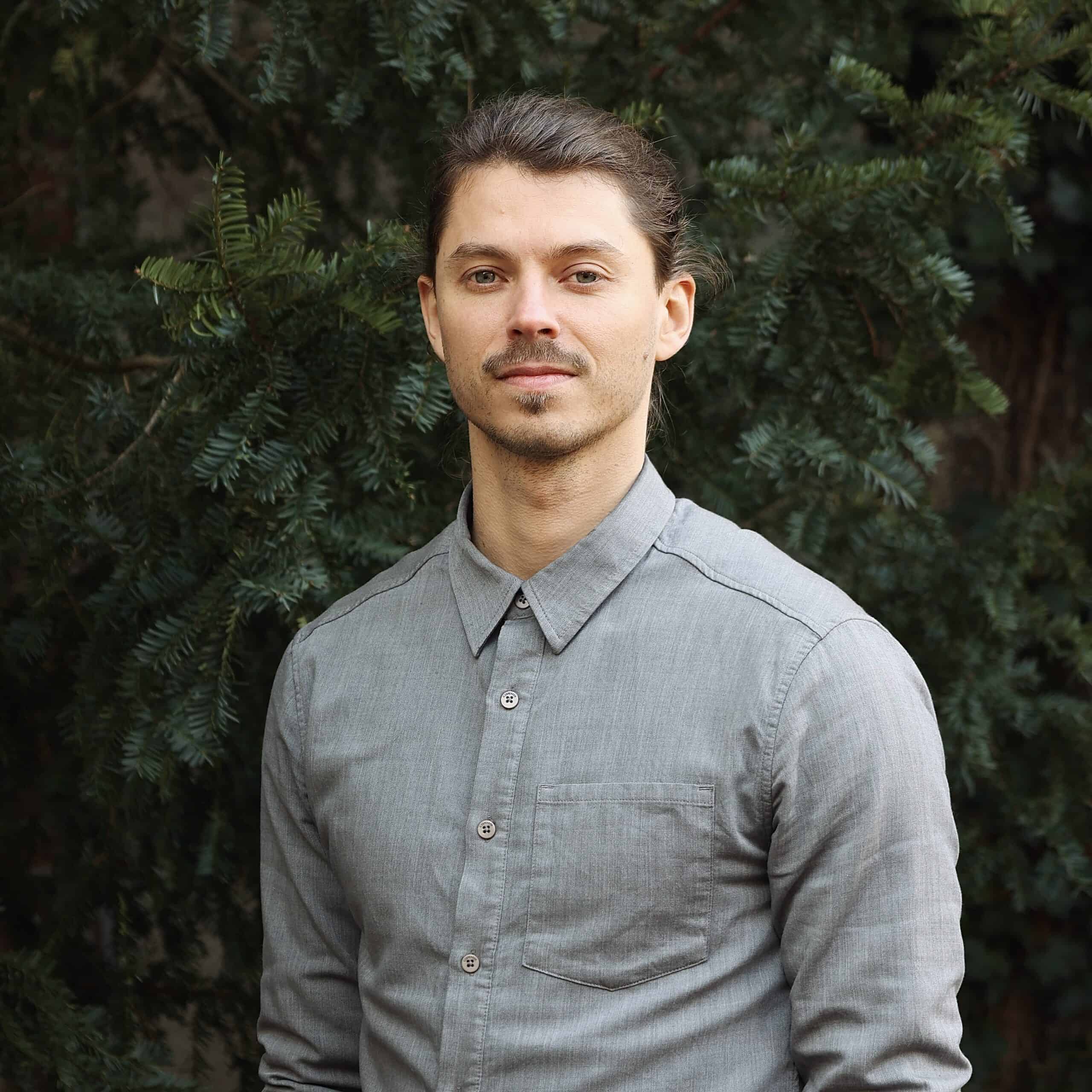 Der Osteopath Christian Kästle steht vor einem grünen Hintergrund in der Natur und schaut etwas ernster in die Kamera