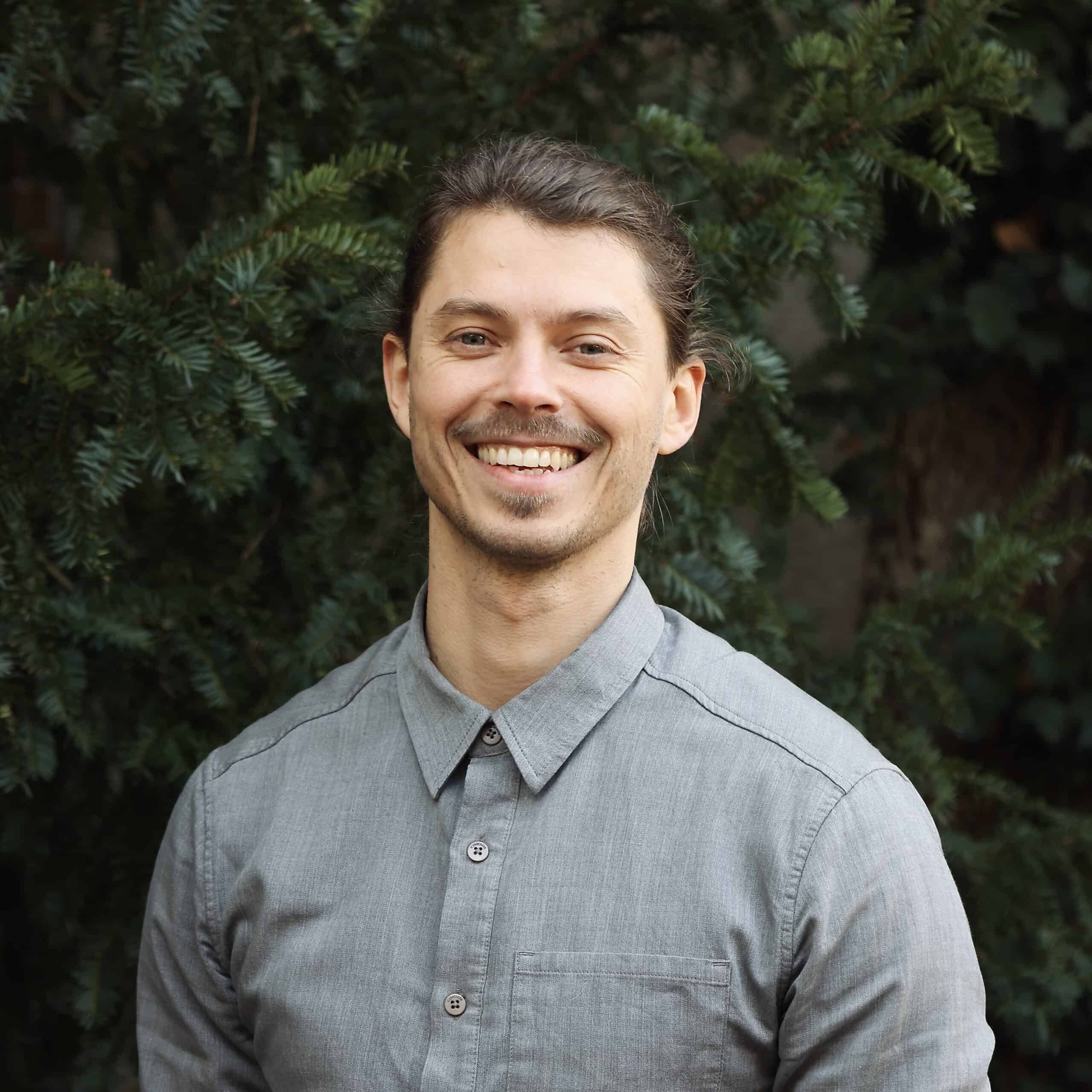 Der Osteopath Christian Kästle steht vor einem grünen Hintergrund in der Natur und lacht in die Kamera